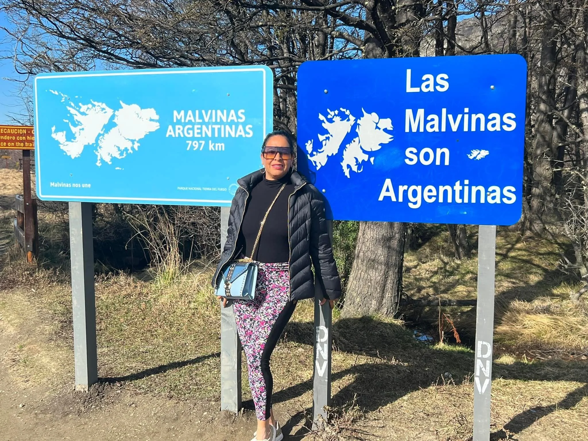 A woman standing in front of two blue signs indicating routes to the Malvinas Islands. One sign is in English and the other in Spanish, with the maps of the islands and distances. The woman is wearing black sunglasses, a black jacket, and floral pants.