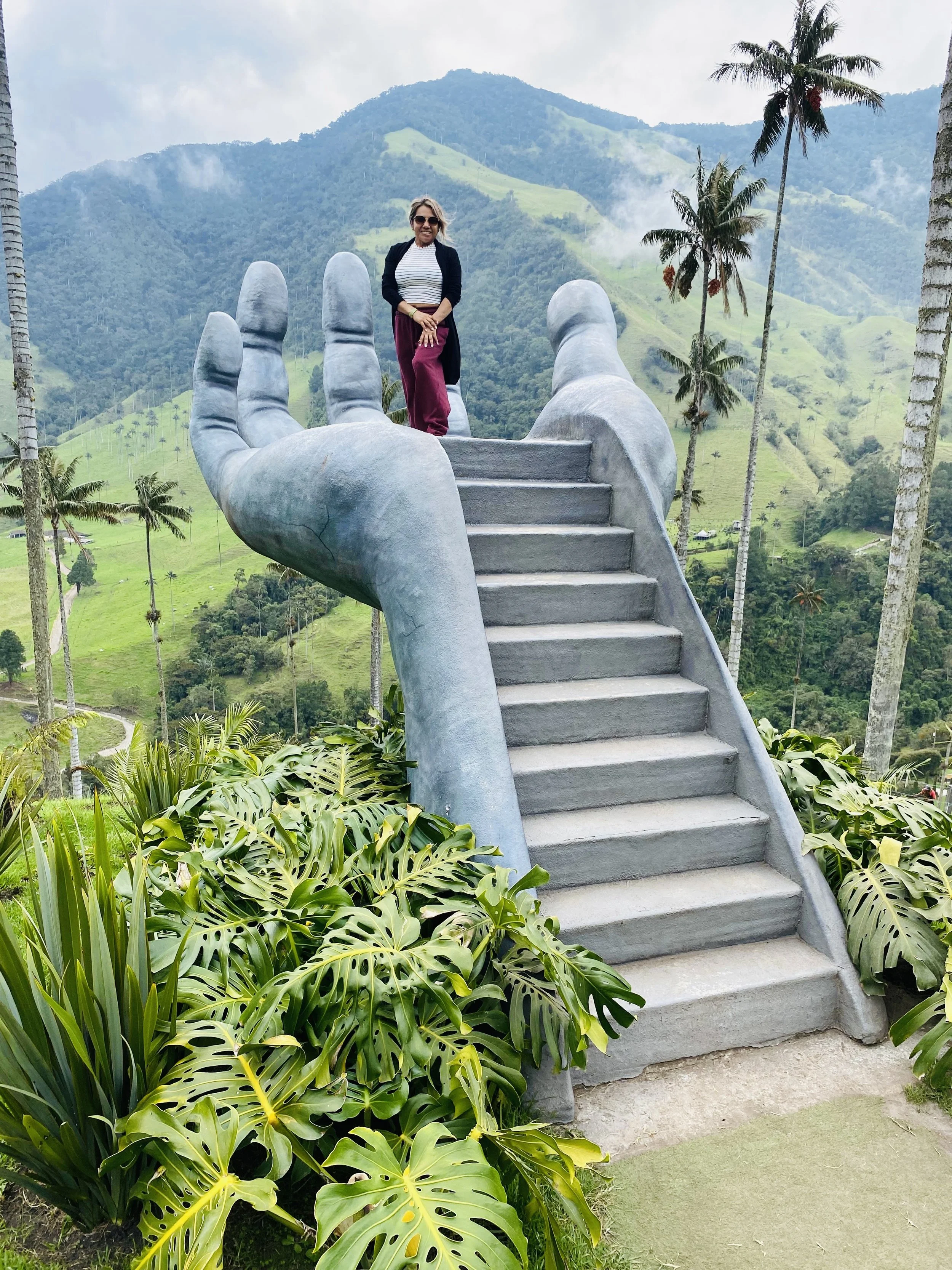 A woman standing on a large, stylized concrete hand that forms part of stairs, with a lush green mountain landscape and tall palm trees in the background.