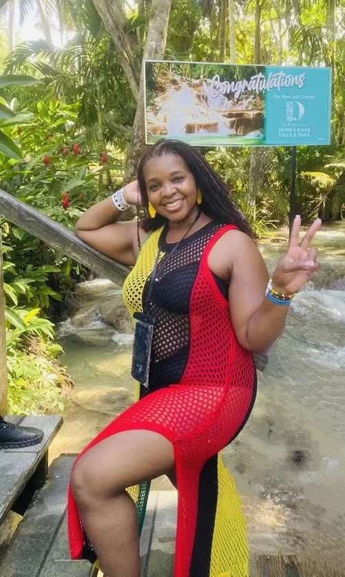 A woman posing cheerfully at a waterfall with a 'Congratulations' sign in the background, wearing a colorful dress and making a peace sign gesture.
