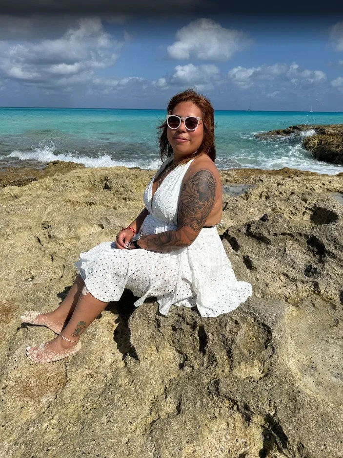 Woman with tattoos wearing sunglasses and a white dress sitting on rocks near the ocean with turquoise water and sailboats in the background.
