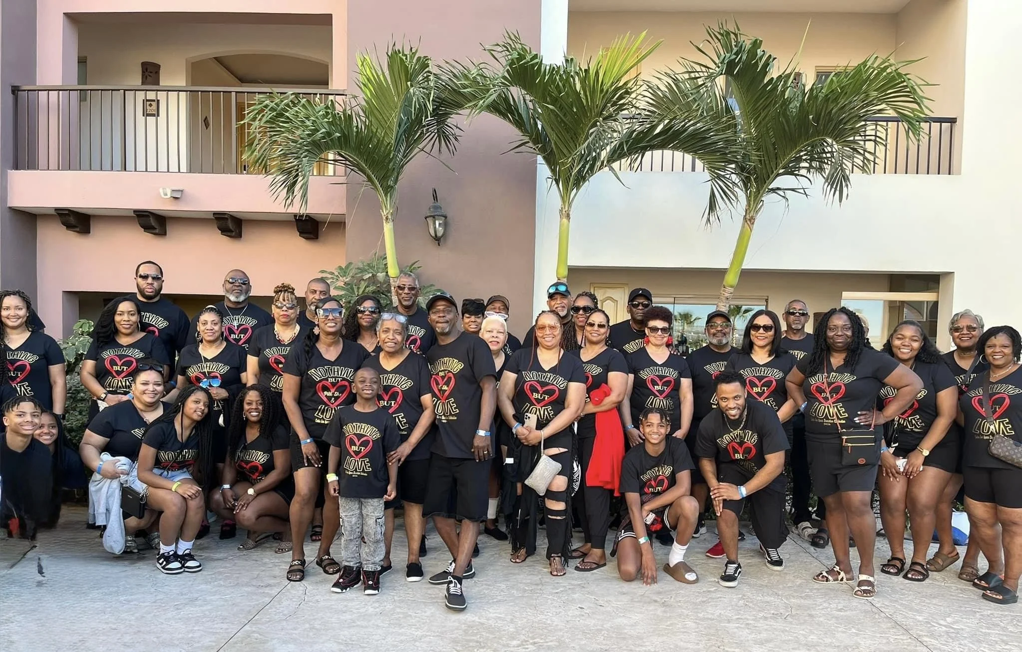 Group of diverse people of different ages wearing matching black T-shirts with a red and white heart logo and the phrase 'Nothing But Love,' standing outside a modern building with pink and beige walls, palm trees, and a concrete pathway.