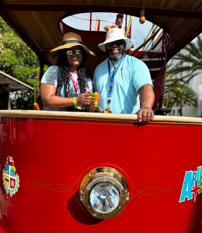 Two people standing inside a red amusement park ride with a wooden roof. The woman on the left is wearing a wide-brimmed straw hat, sunglasses, a gray T-shirt, and a red lanyard. The man on the right is wearing a white hat, sunglasses, a light blue T-shirt, and a blue lanyard. They are smiling and holding onto a control lever or handle. In the background, there is a spiral staircase and a woman standing at the top of the ride with trees and a cloudy sky visible behind.