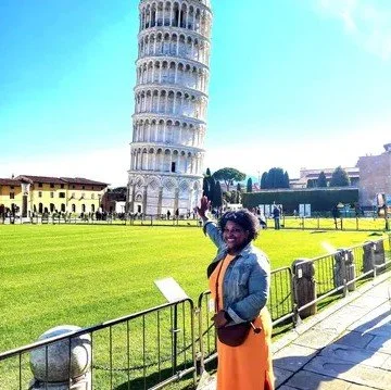 A woman smiling and pointing at the Leaning Tower of Pisa in Italy, with a grassy area and historic buildings in the background.