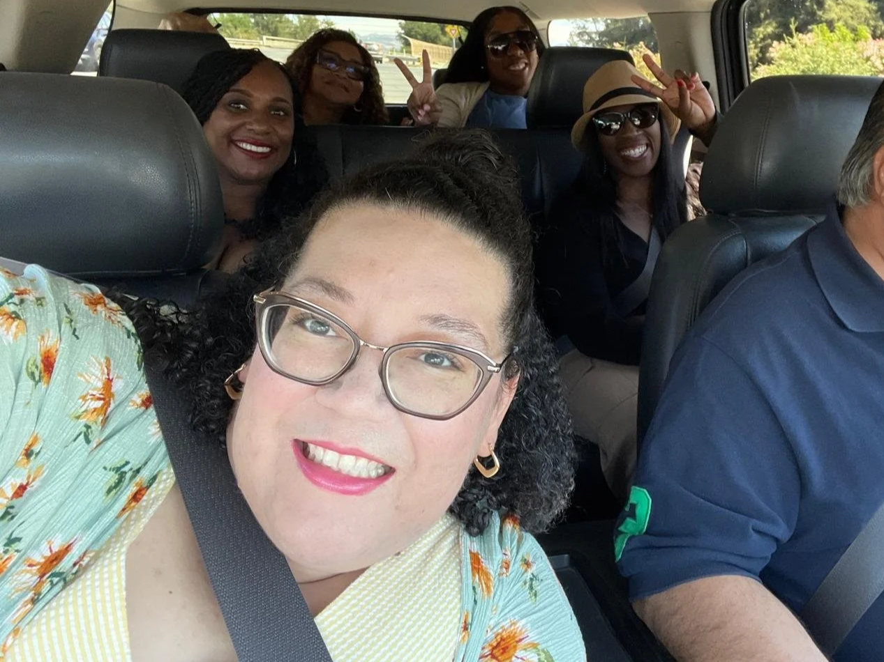 A group of six women and one man inside a car, smiling and making peace signs. The woman in the front is taking a selfie, wearing glasses and a floral top. The others are seated behind her, some wearing sunglasses, and one woman is wearing a hat. They appear cheerful and enjoying their time together.