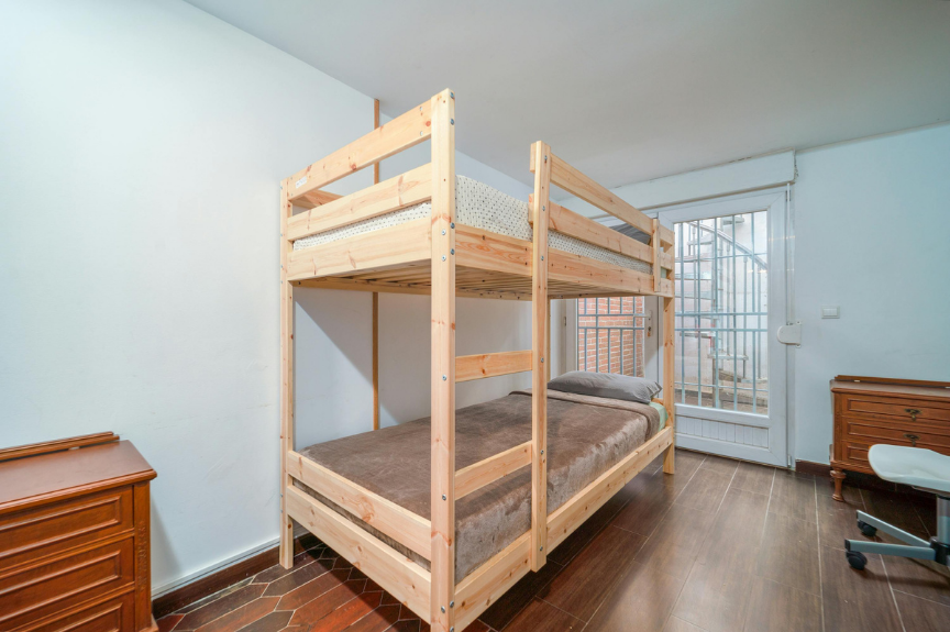 Room with a wooden bunk bed, a window with metal bars, a wooden side table, and a white office chair.