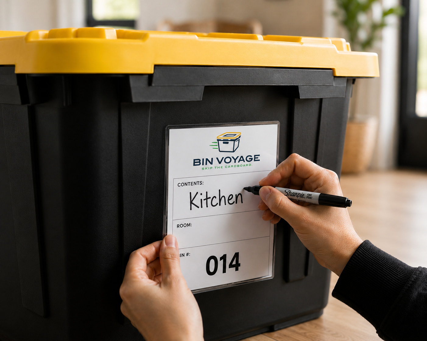 Person writing 'Kitchen' on a label attached to a black and yellow bin labeled 'Bin Voyage' with the slogan 'Skip the Cardboard.'