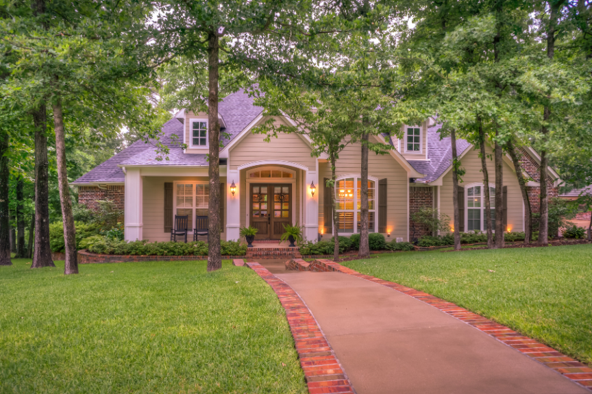 Front porch of a suburban house with a brick pathway, surrounded by greenery and mature trees, during dusk with warm exterior lighting.