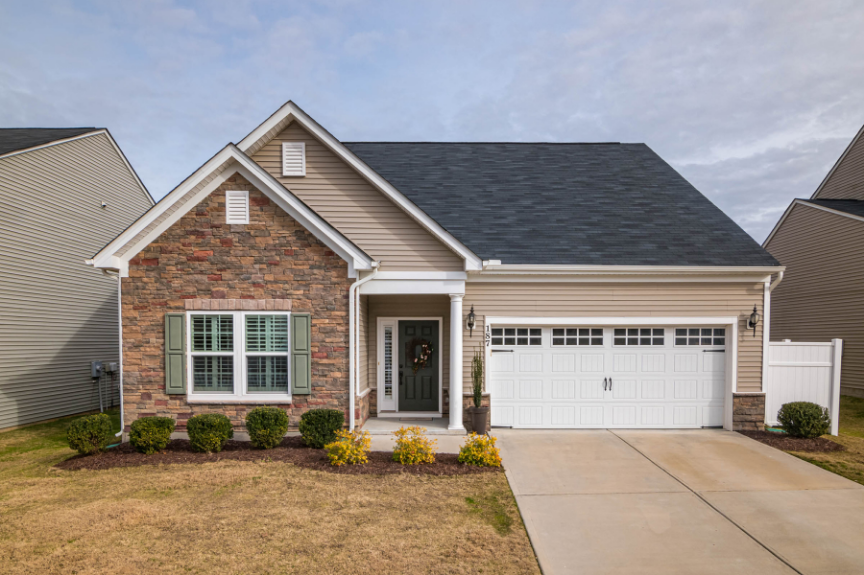 Front view of a modern suburban house with beige siding, brick facade, a dark gray roof, white garage door, and a small front porch decorated with a wreath and potted plants. There are manicured bushes and a concrete driveway.