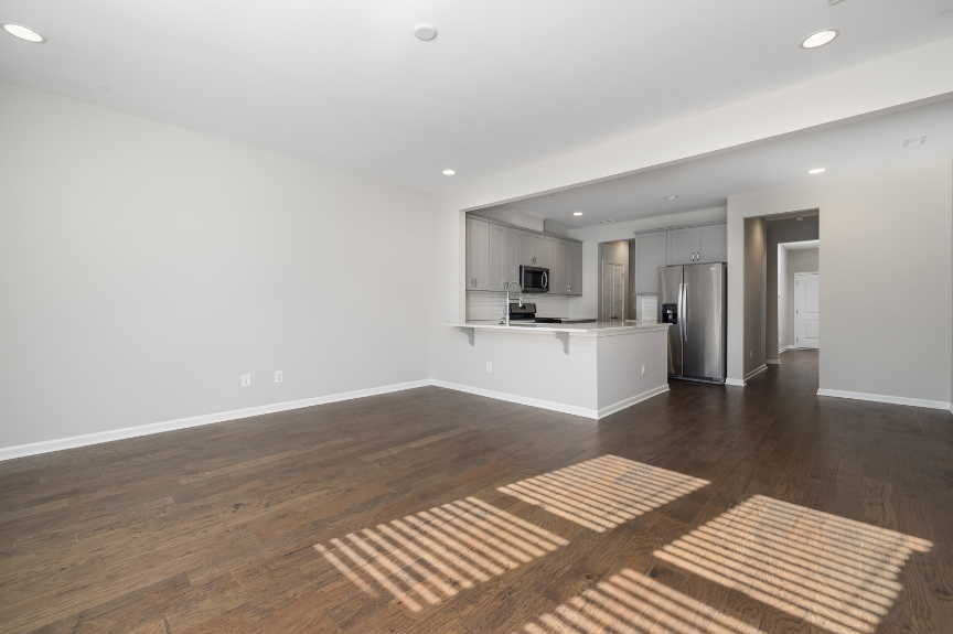 Empty living room with hardwood floors, white walls, and sunlight coming through window blinds, showing part of a kitchen with stainless steel appliances.
