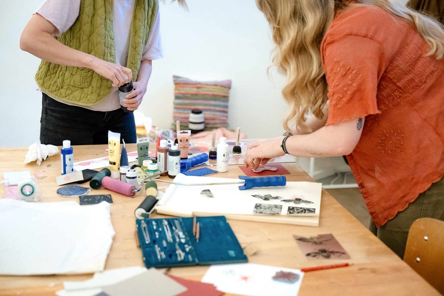 Zwei Frauen arbeiten an einem Tisch mit Tinten, Papieren, Pinseln und Druckmaterialien, möglicherweise beim Basteln oder Drucken.