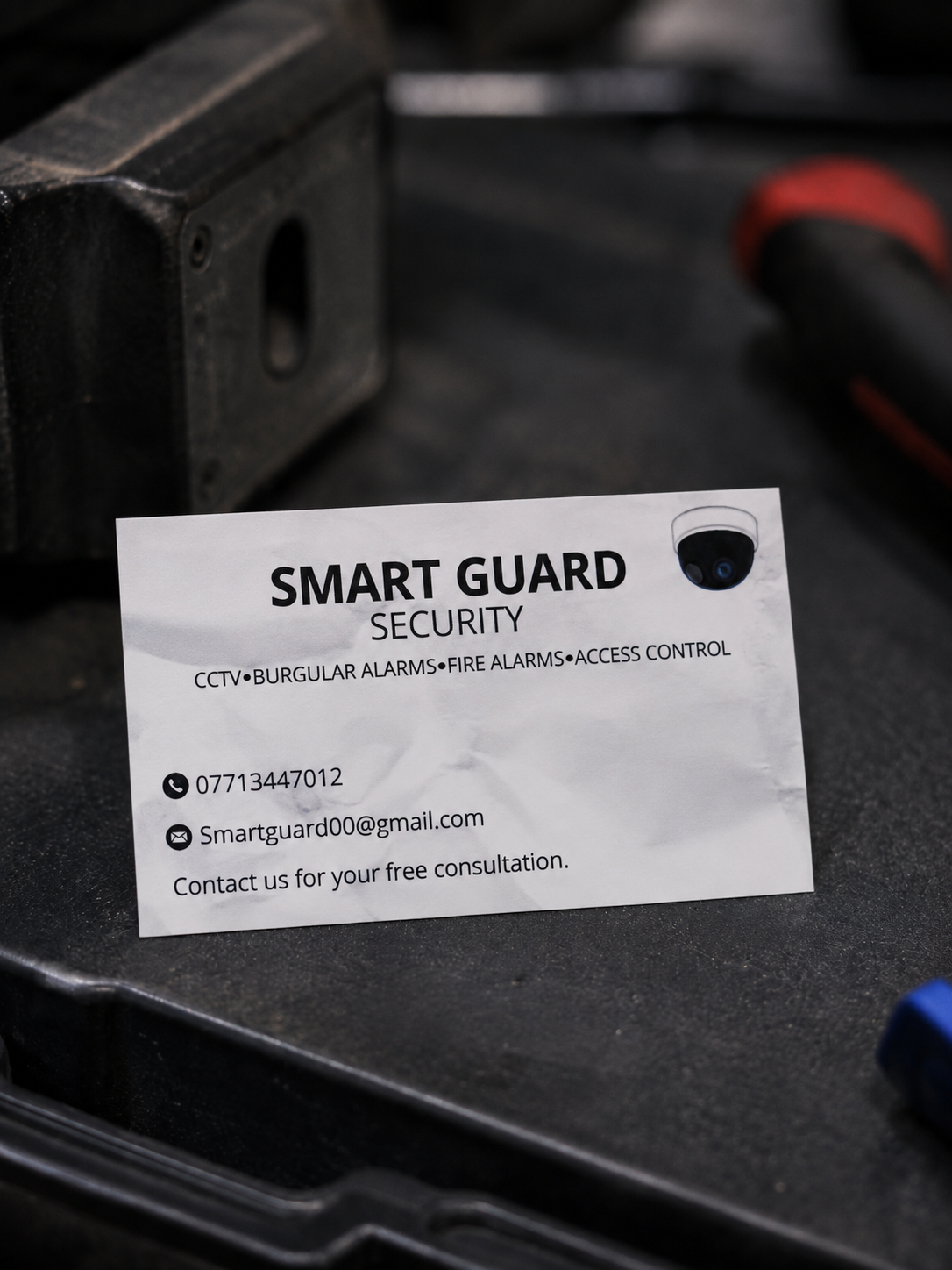 Business card for Smart Guard Security company, featuring contact information and listing services like CCTV, burglar alarms, fire alarms, and access control. The card is placed on a black surface with tools and equipment nearby.