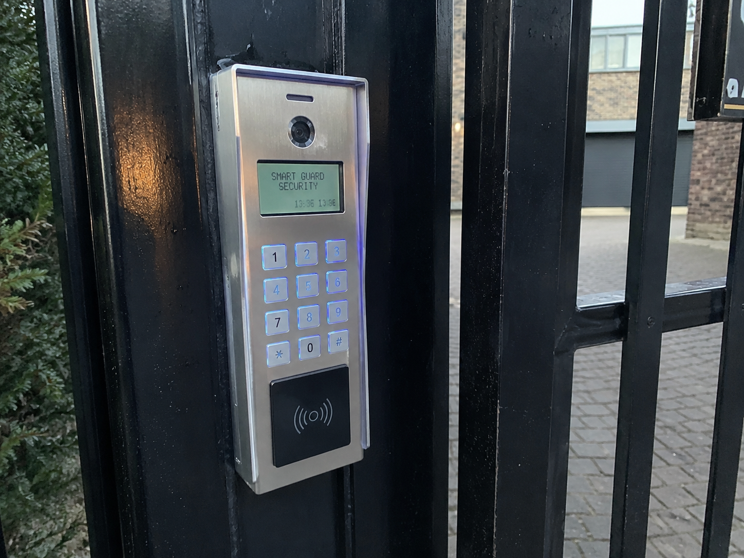 A digital gate access control panel mounted on a black metal gate. The panel has a keypad with numbers and symbols, a small screen displaying 'SMART GUARD SECURITY,' a camera lens above the screen, and an RFID reader at the bottom. The background shows a paved area and a building with brick walls and windows.