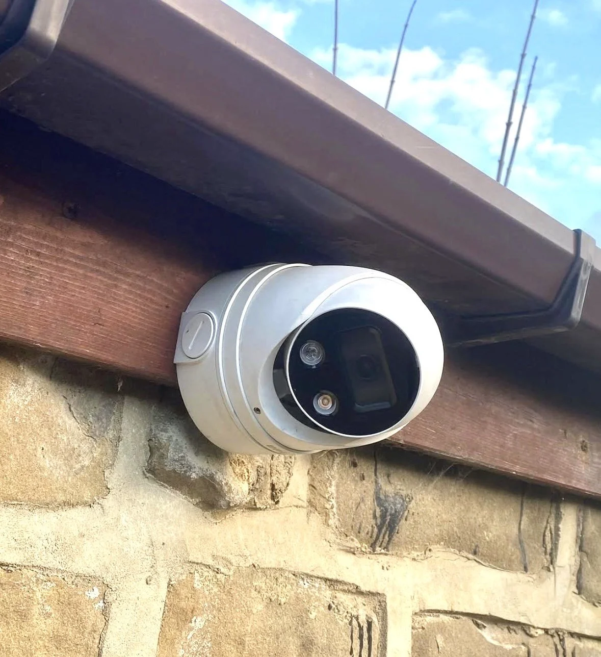 Security camera mounted under the eave of a stone building with a brown gutter and clear sky in the background.
