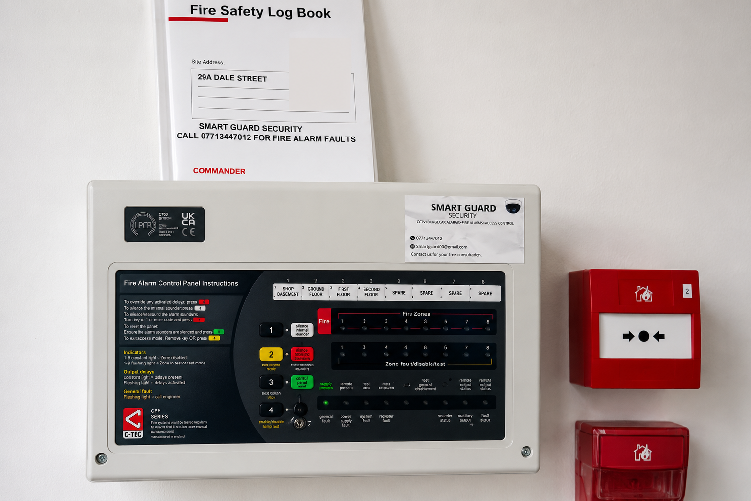 A fire alarm control panel mounted on a white wall with instructions, a smart guard security sticker, and two red fire alarm call points nearby.