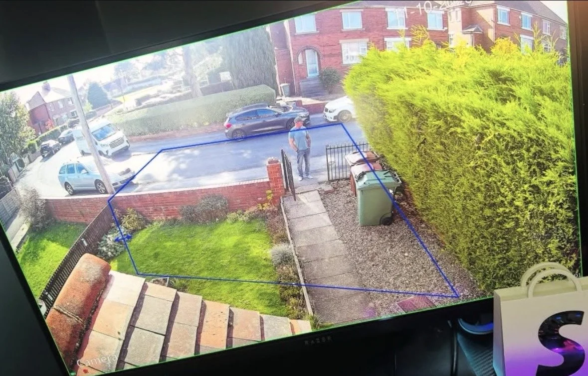 View from a security camera showing a person walking on the sidewalk on a residential street, with parked cars, a brick wall, and a large green hedge.