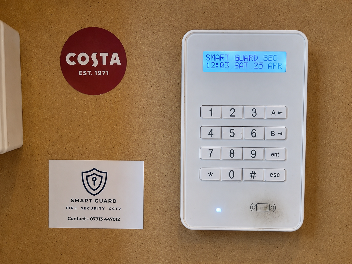 A keypad security system with a blue display screen showing 'SMART GUARD SEC 12:03 SAT 25 APR'. To the left, a round red sign with white text reading 'COSTA EST. 1971'. Below the sign, a white business card for 'SMART GUARD FIRE SECURITY CCTV' with a phone number. The keypad is on a brown surface.