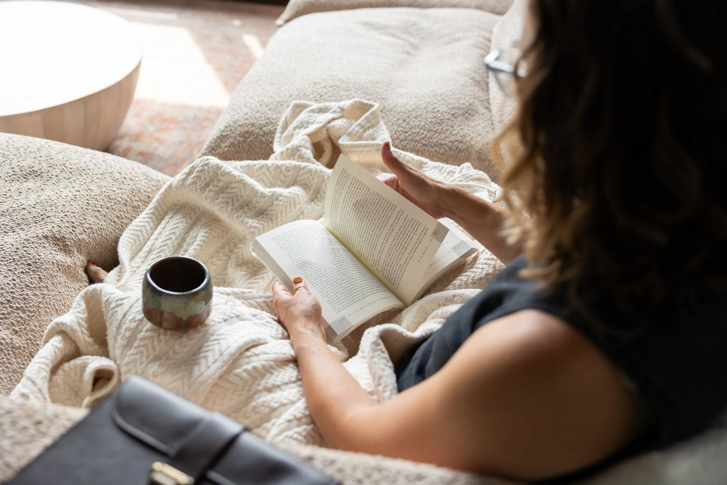A person sitting on a beige couch reading a book, with a cup of coffee or tea nearby.