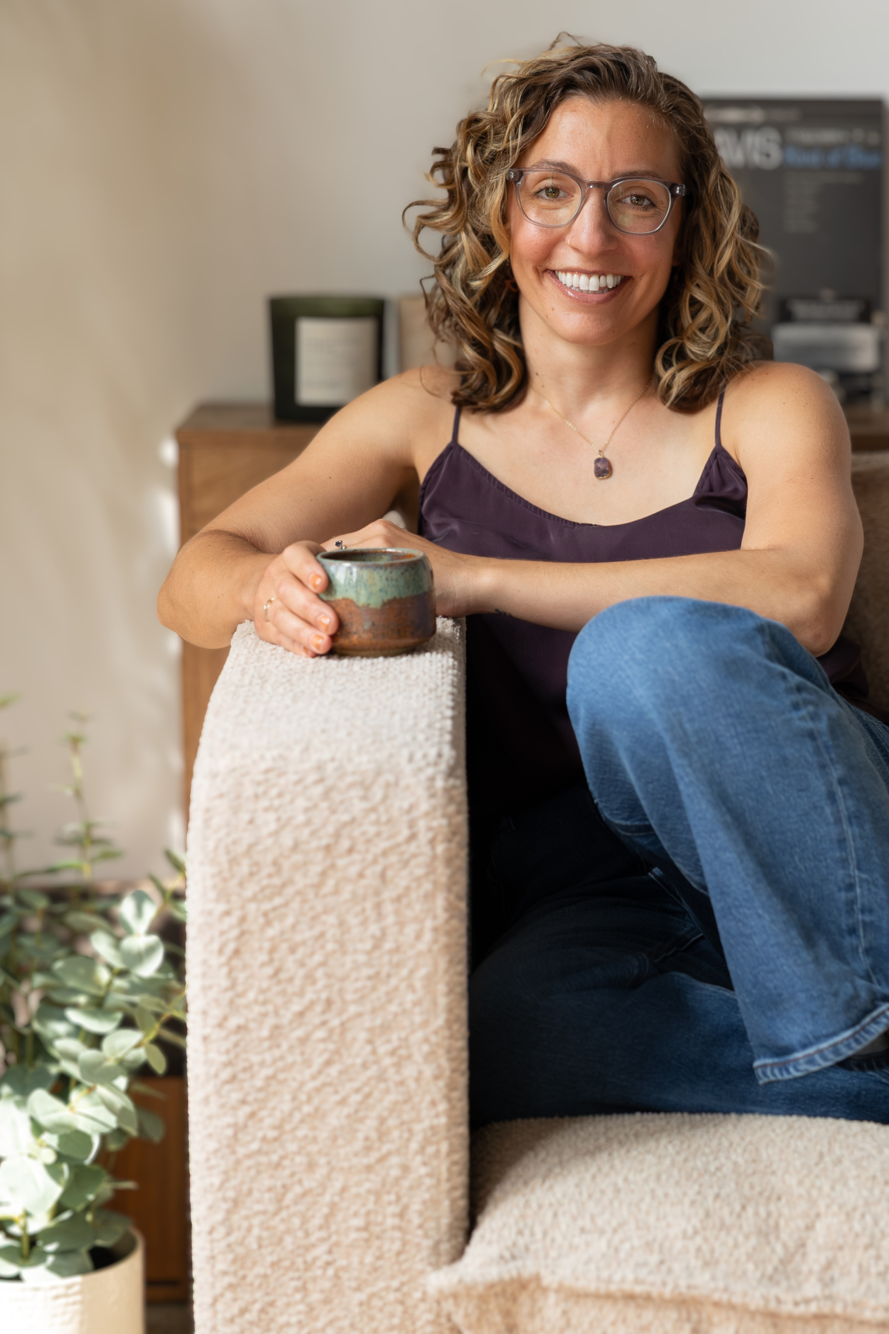 A woman with curly hair and glasses smiling while sitting on a beige textured couch. She is wearing a purple spaghetti strap top and holding a ceramic mug. In the background, there is a wooden cabinet with a plant on the side and a blackboard with writing.