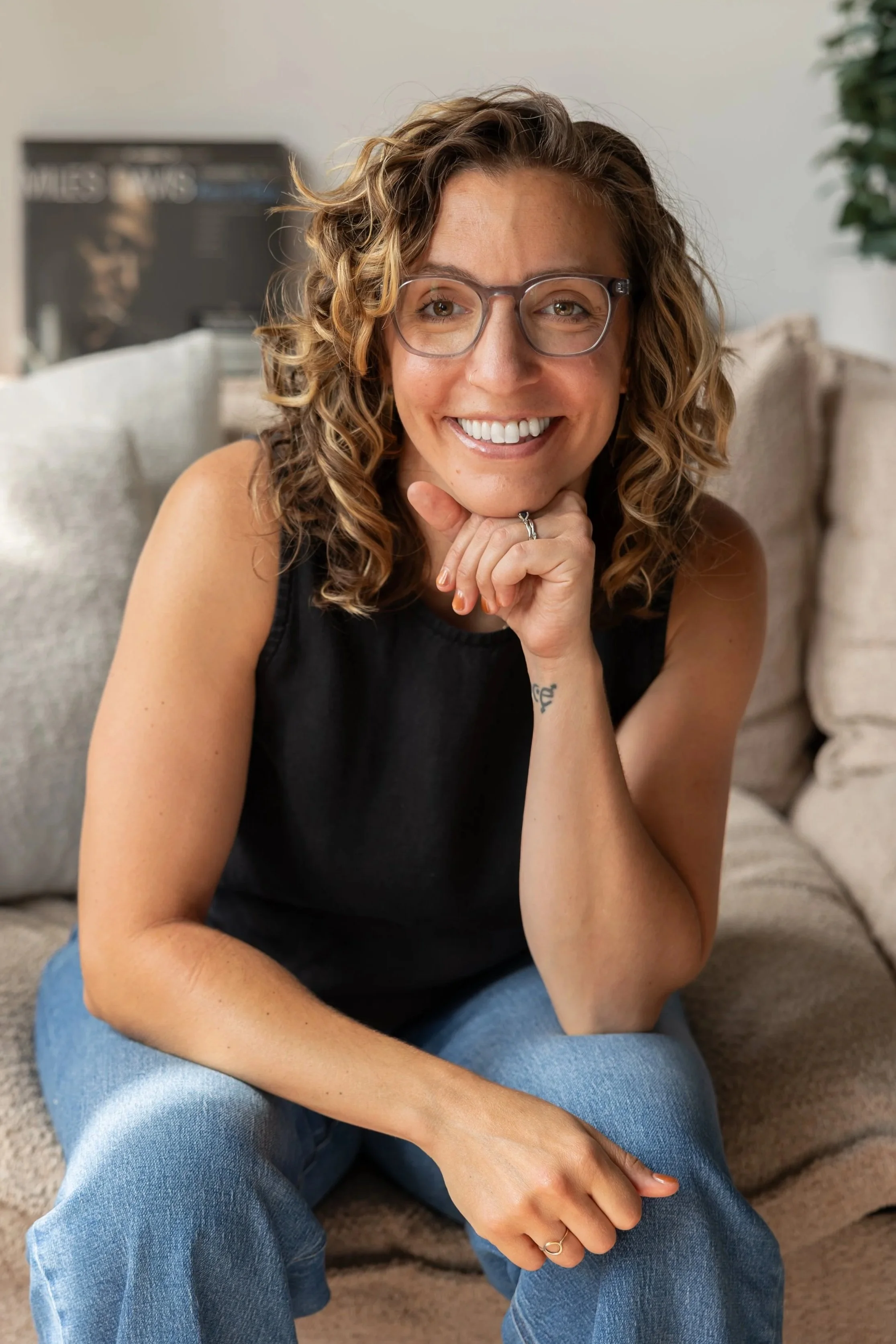 A woman with curly hair and glasses sitting on a beige couch, smiling, wearing a black sleeveless top and blue jeans, resting her chin on her hand.