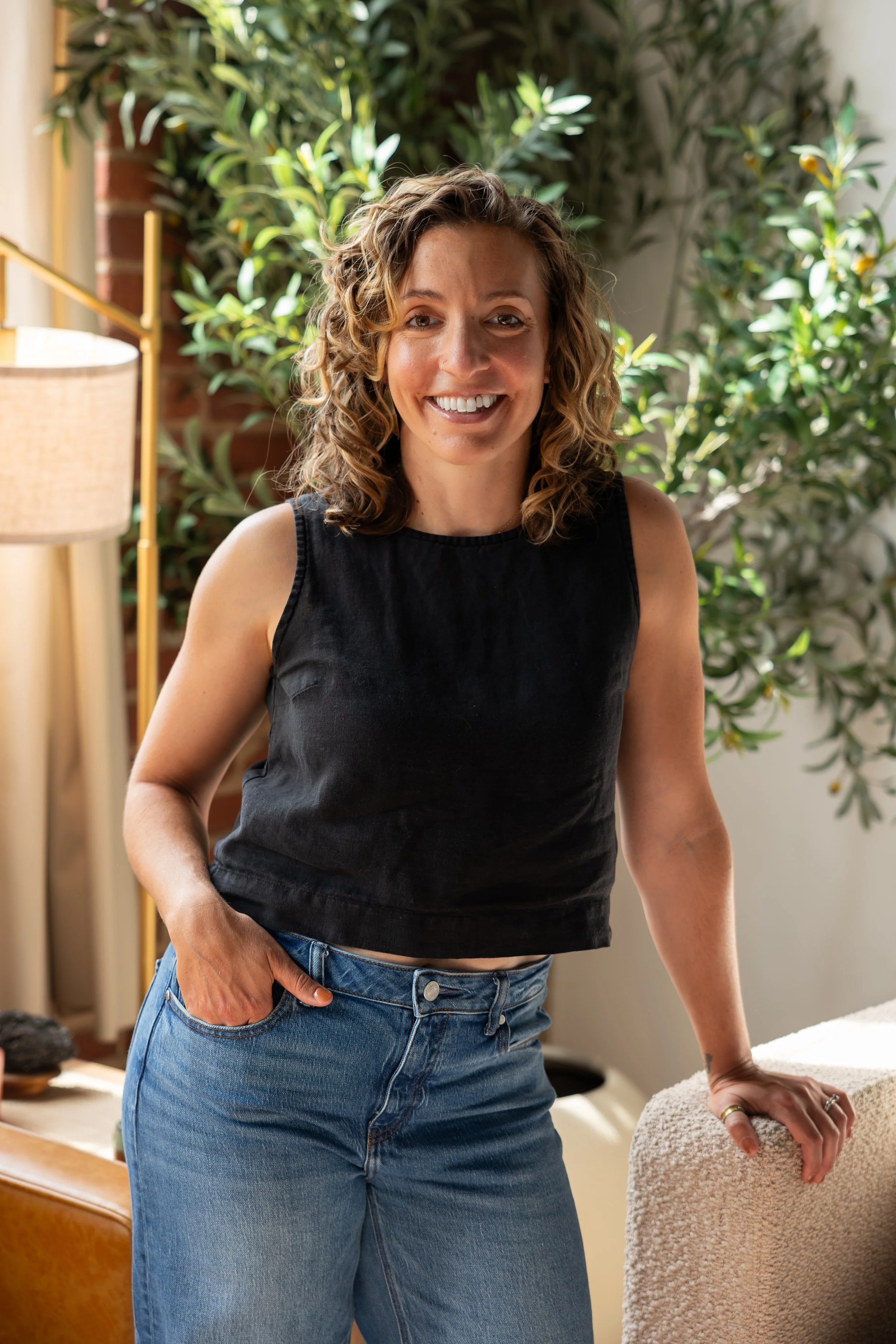 A woman with curly hair wearing a sleeveless black top and blue jeans, smiling in a room with green plants and a table.