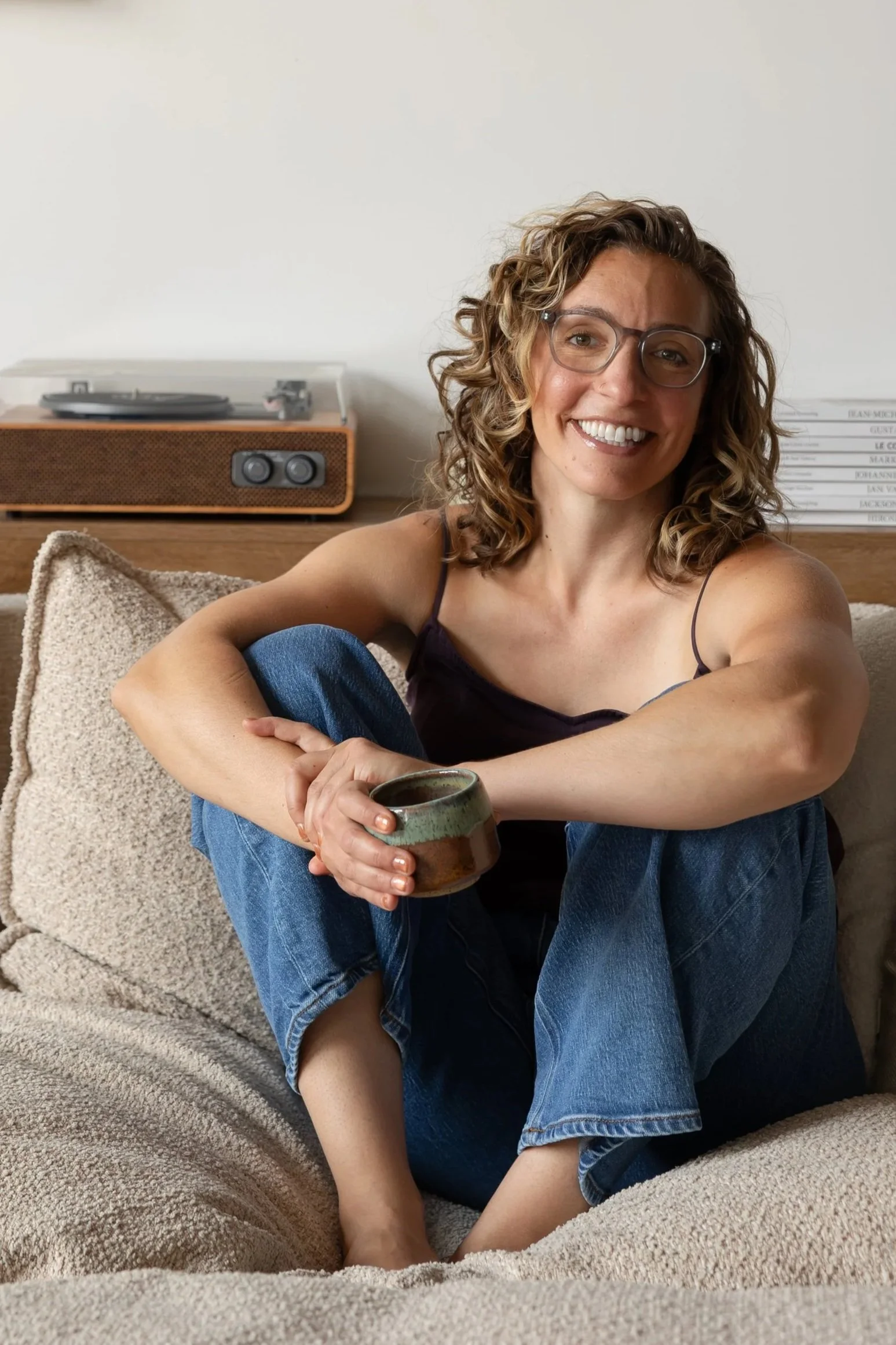 A woman with curly hair and glasses sitting on a beige couch, smiling, holding a mug, with a record player on a wooden table behind her.