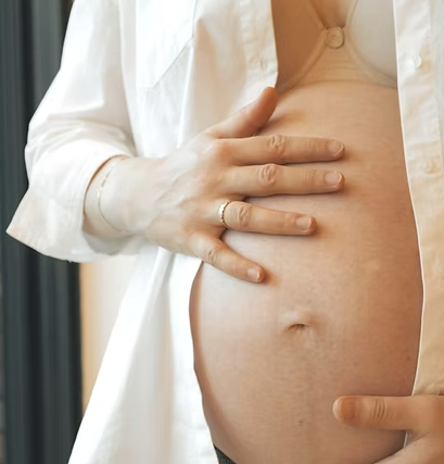 Close-up of a pregnant woman's belly with a hand gently resting on it, wearing a white open shirt