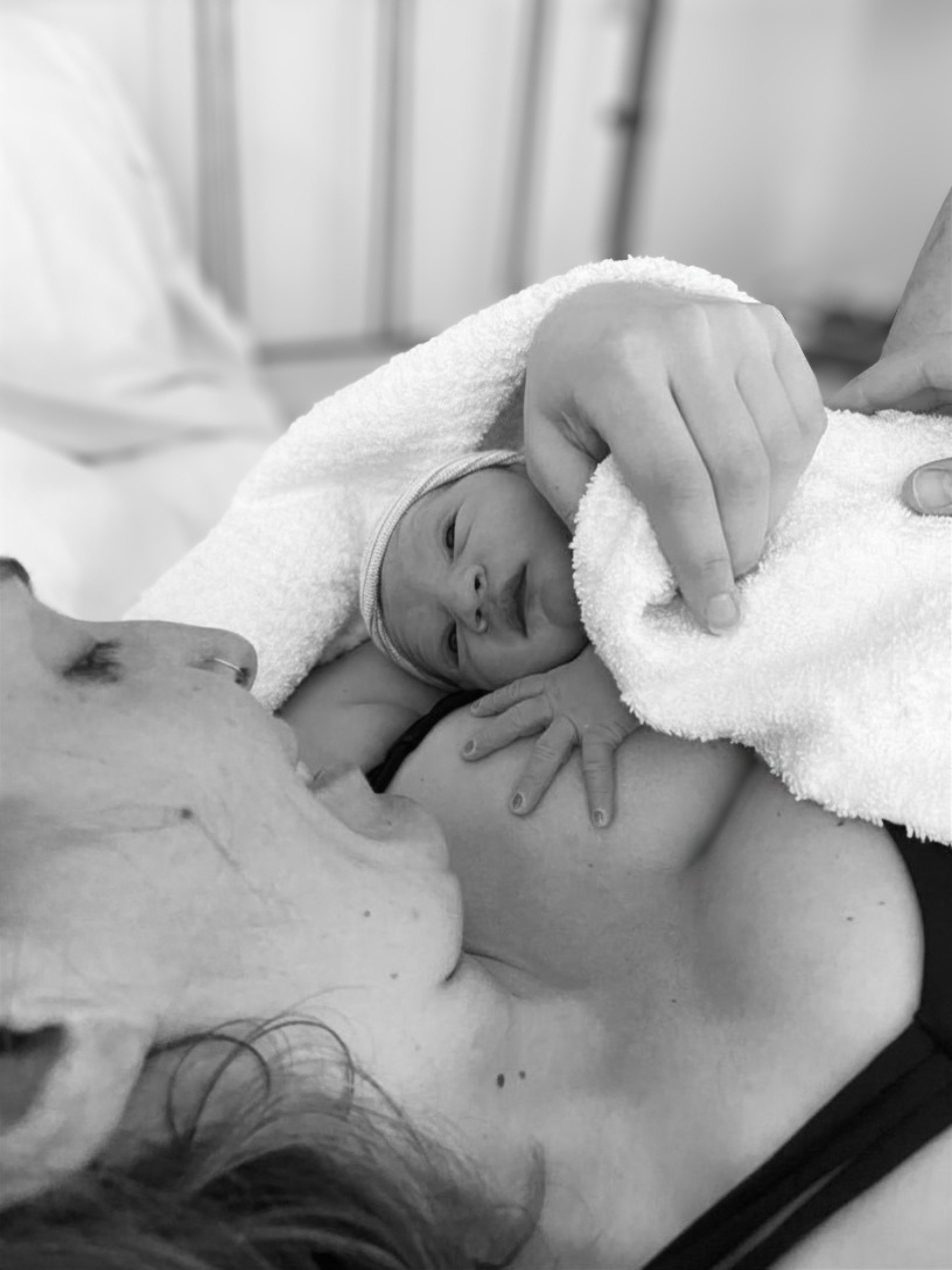 Close-up black and white photograph of a newborn baby lying on a towel, with a person gently holding the baby's hand, in a medical setting.