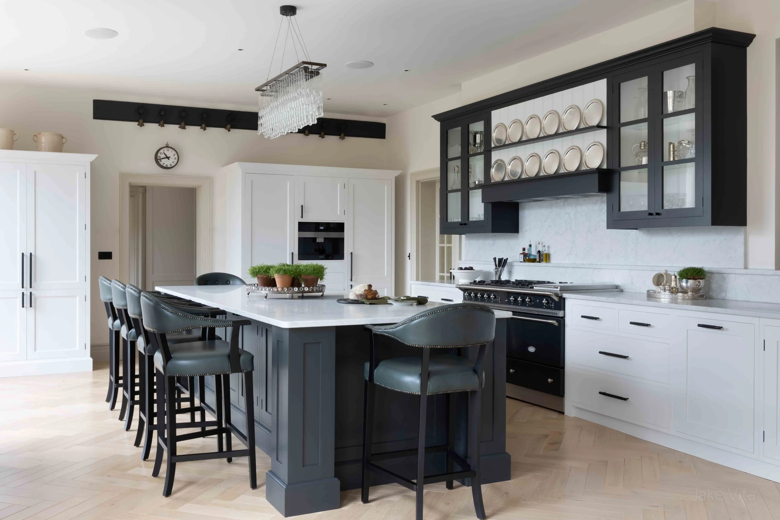 A modern kitchen with white and black cabinetry, a large island with black chairs, a chandelier, and decorative plates displayed on a black wall unit.
