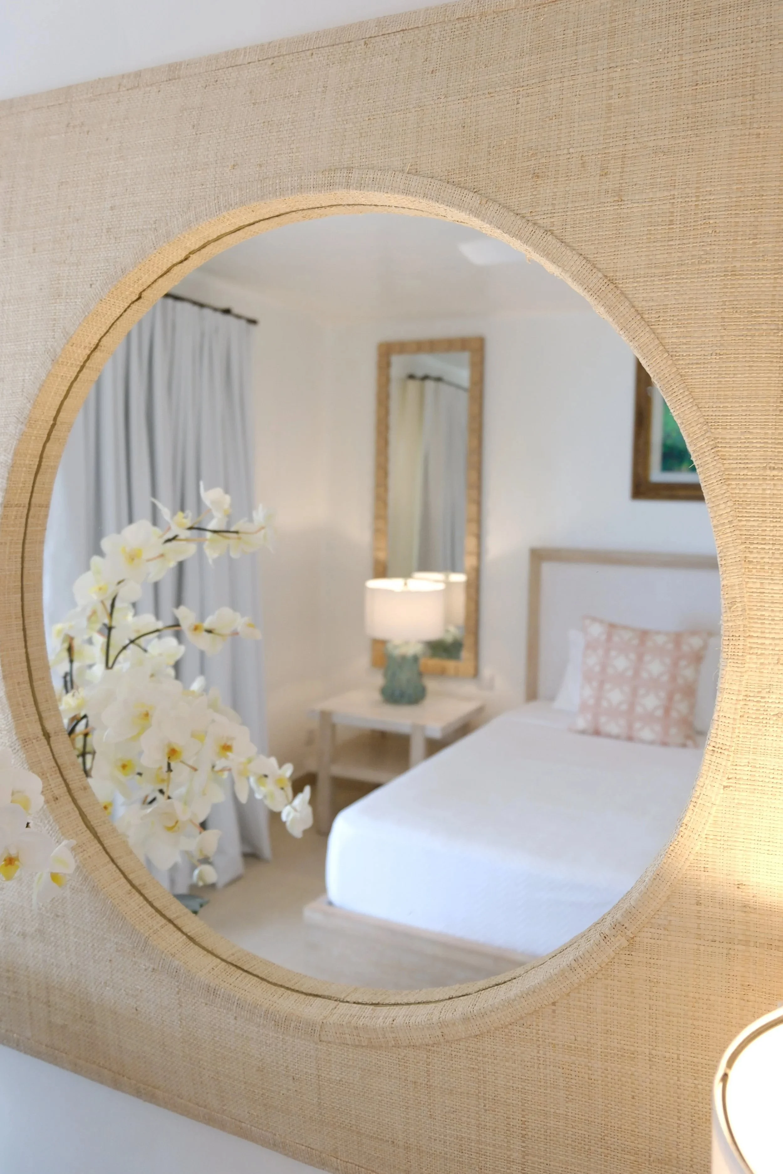 A room with a bed, a nightstand with a lamp, a framed mirror on the wall, light curtains, a floral arrangement, and a window visible through a circular cutout in beige textured fabric.