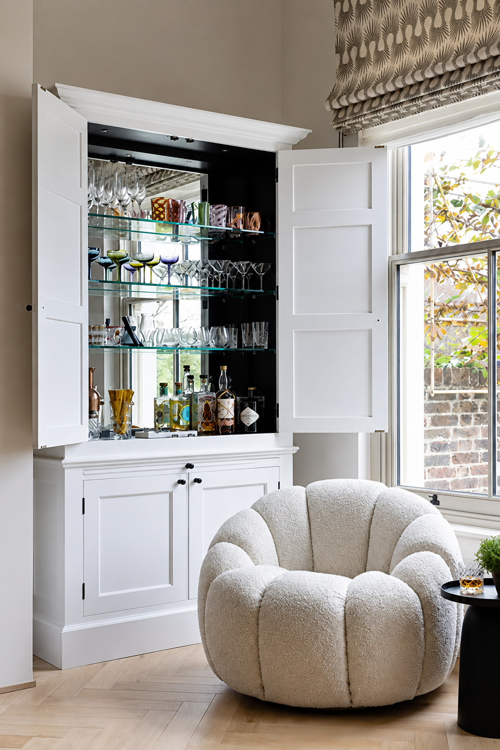 A cozy room corner featuring a white cabinet with glassware and liquor bottles inside, an open window with a view of a brick wall and greenery, and a plush, rounded cream-colored armchair.