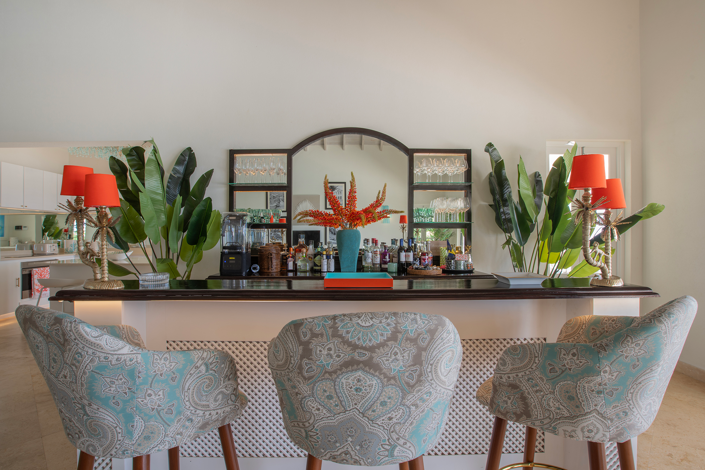 A kitchen or bar area with a large mirror above a wooden counter. The counter has bottles of alcohol and glassware, with two red lamps on either side. There's a large blue vase with orange and red flowers in the center. The background features tall green plants on either side of the mirror and patterned upholstered chairs around a table in the foreground.