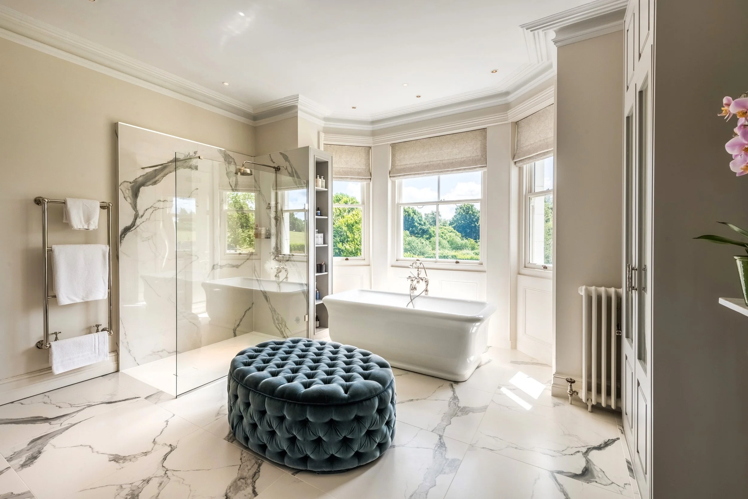 A luxurious bathroom with large windows, a freestanding bathtub, a glass shower enclosure with marble walls, a marble-topped vanity, and a tufted ottoman. Natural light floods the space, highlighting the elegant decor and countryside view outside.
