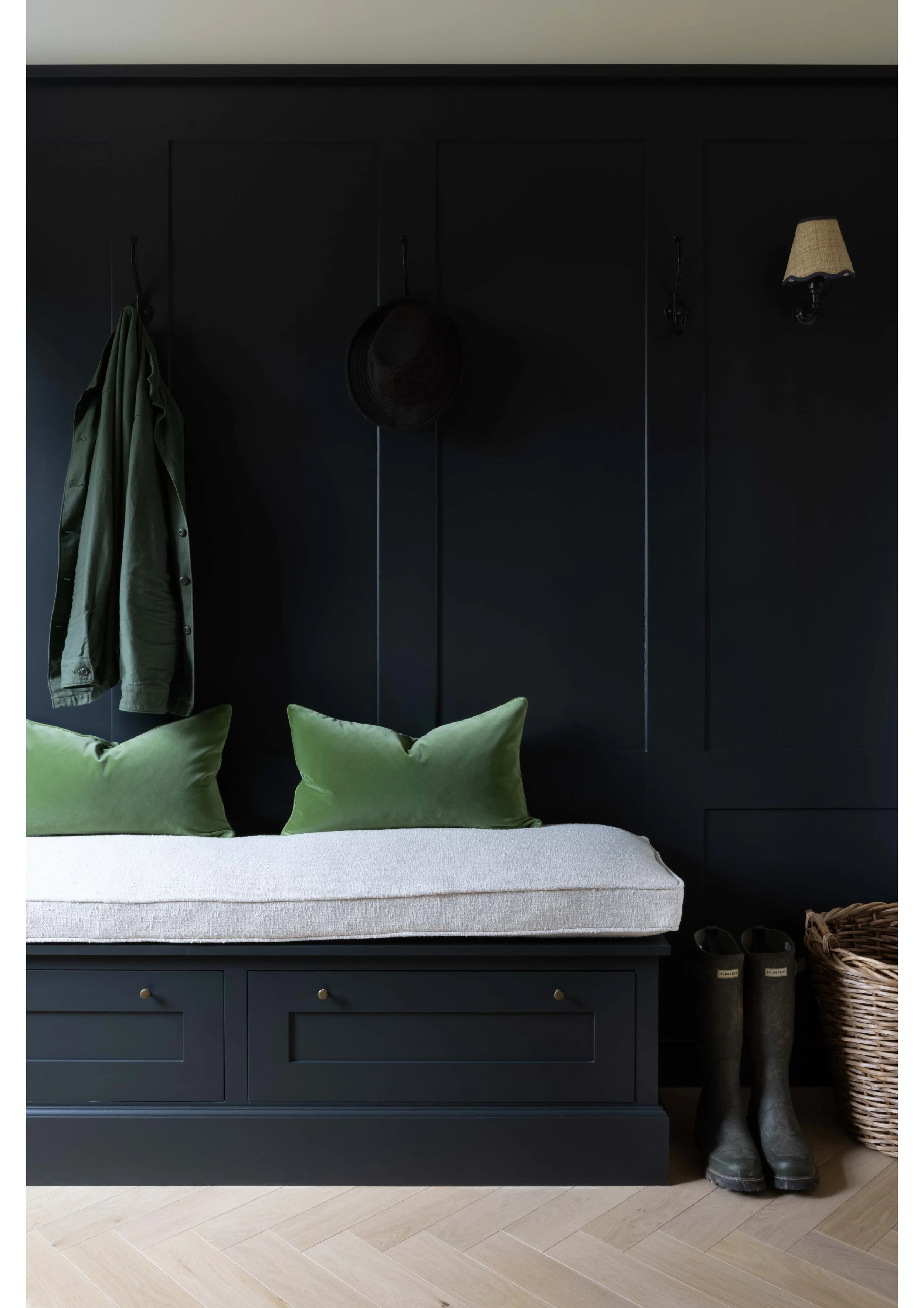 A bedroom with a dark blue built-in bed frame and storage drawers, a white mattress, three green pillows, a black headboard with hooks holding a coat and a hat, two rain boots, a woven basket, and a shoe rack on a light-colored wood floor.