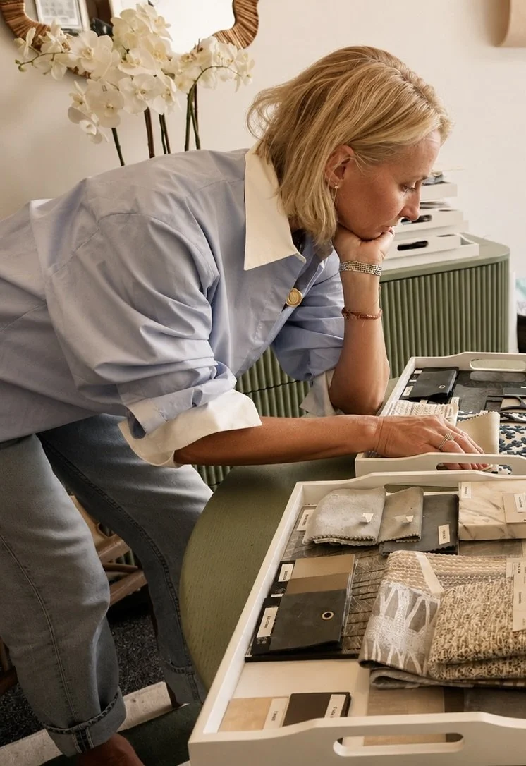 A blonde woman leaning on a table, looking at fabric swatches and samples, with a white orchid plant in the background.