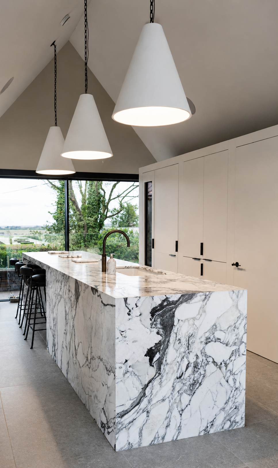 Modern kitchen with a marble island, black stools, white cabinets, pendant lights, and large windows showing greenery outside.