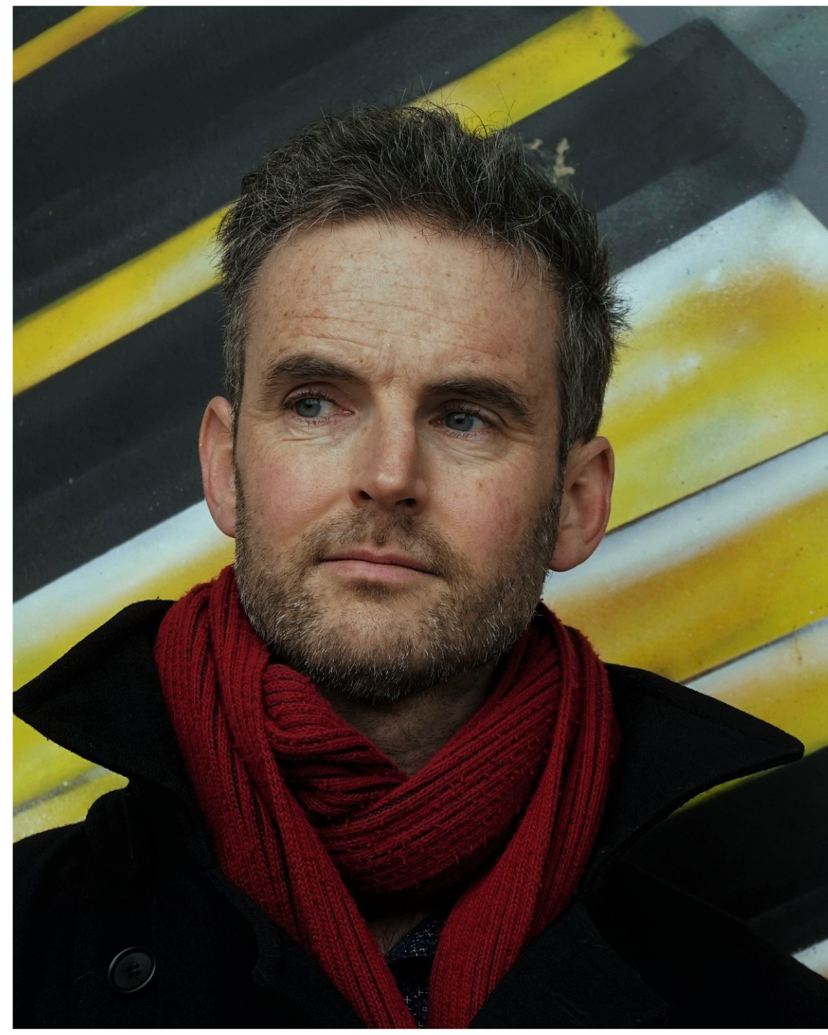 Close-up of a man with short dark hair, beard, and blue eyes, wearing a red scarf and black coat, standing in front of a yellow and black striped background.