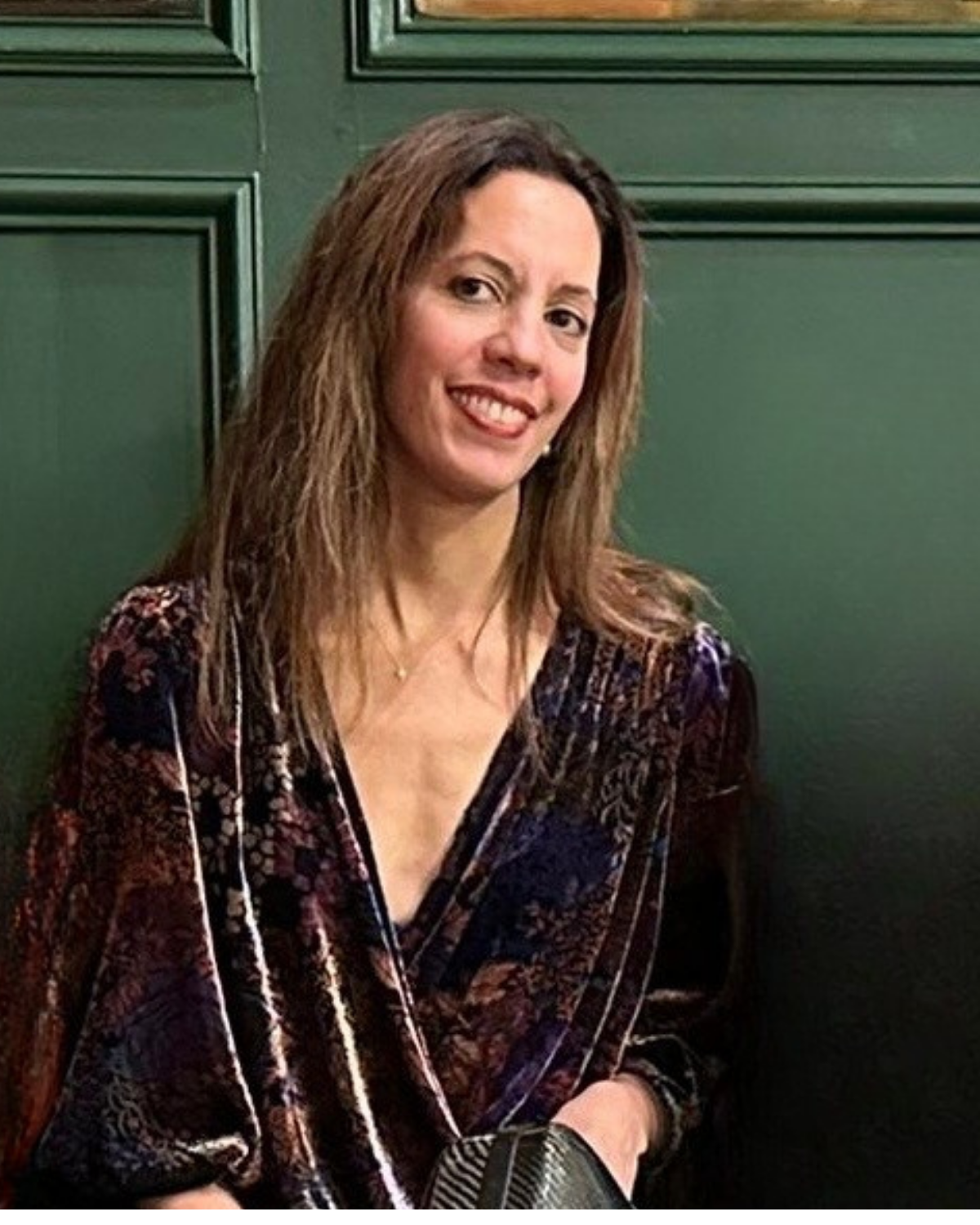 A woman with shoulder-length brown hair smiling, wearing a dark patterned top, sitting in front of green paneled walls.