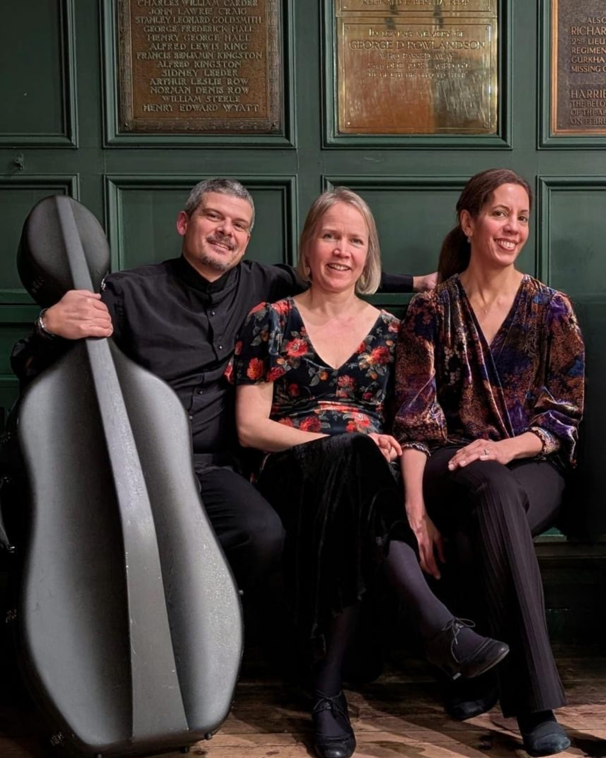 Three people sitting on a green wall bench with bronze plaques in the background. The person on the left is holding a large gray suitcase and wears a black shirt, the middle person wears a floral top, and the right person wears a patterned blouse. They are smiling and posed for the photo.