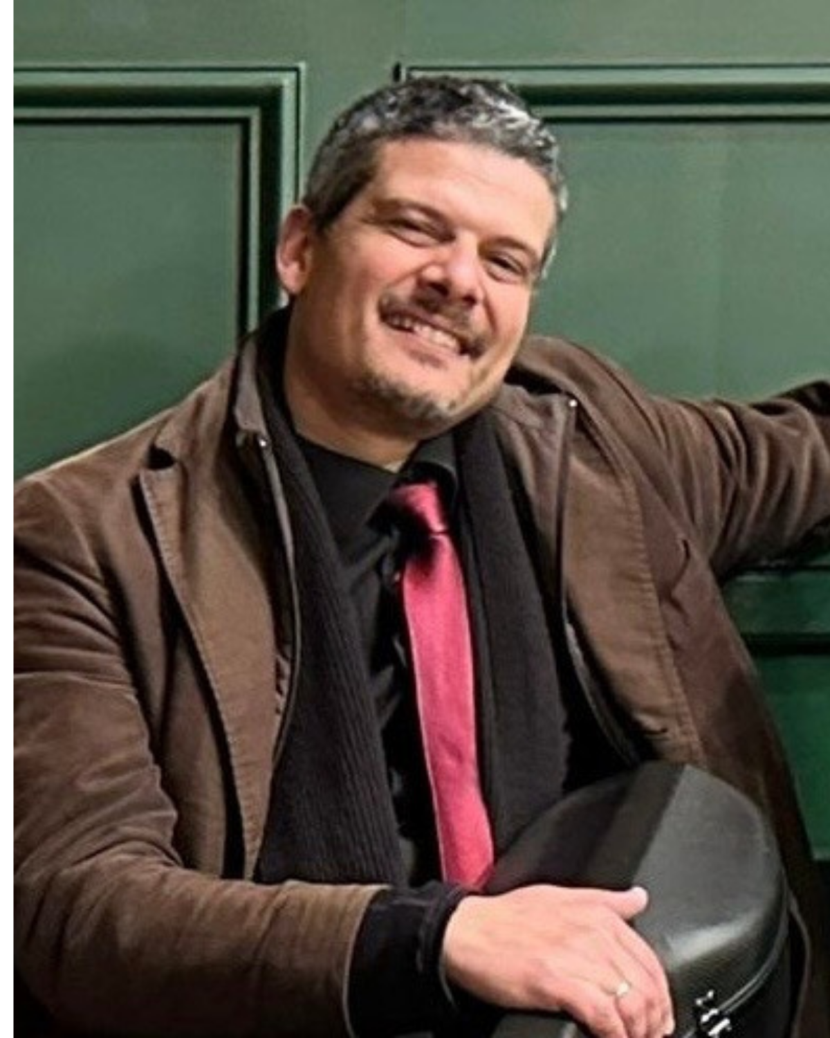 Man smiling, sitting against green wall, wearing a brown jacket, black shirt, and pink tie, with a fedora hat in his hand.