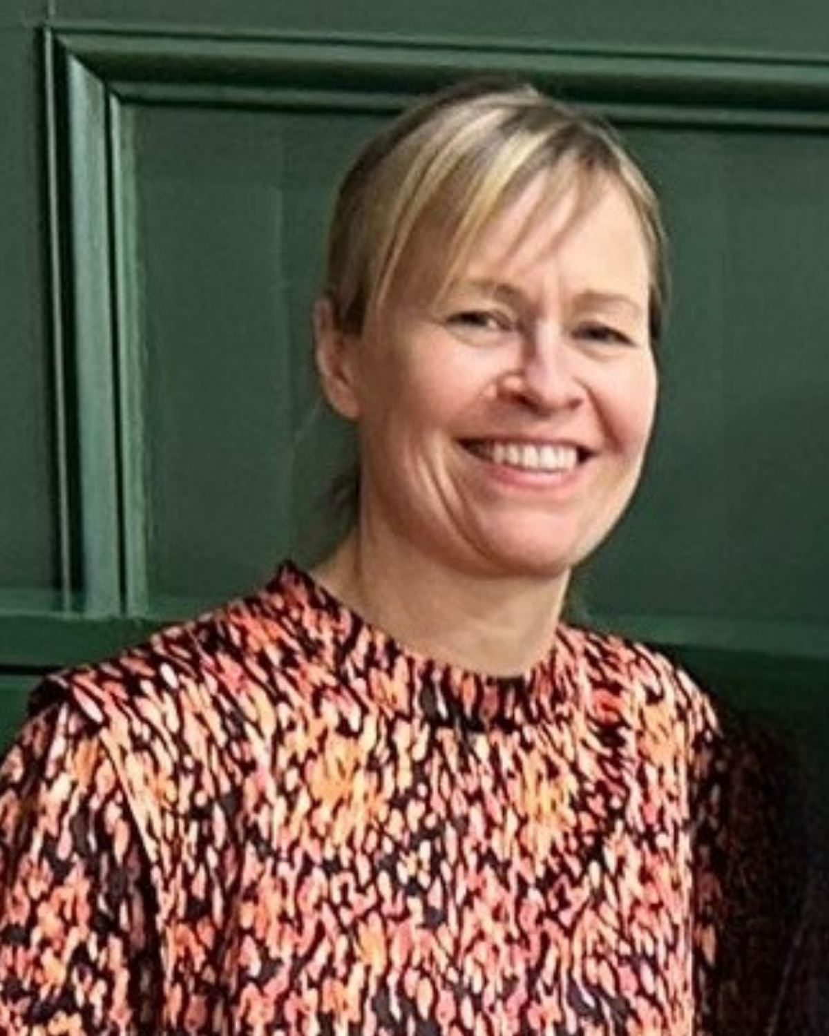 A woman with short blonde hair smiling, wearing a patterned blouse, standing in front of a green cabinet.