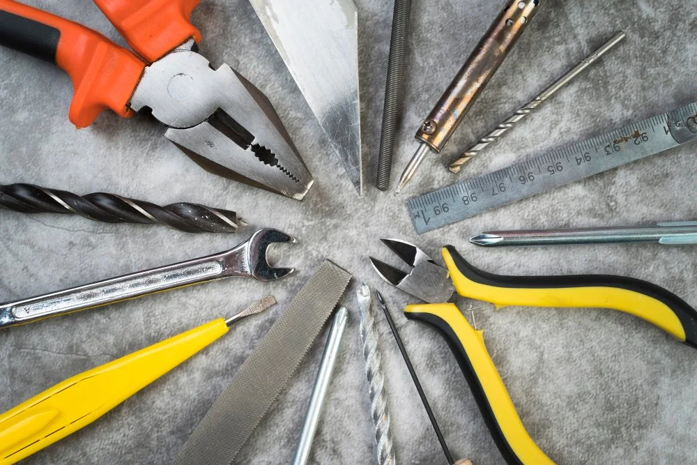 A collection of various hand tools arranged in a circle on a surface, including pliers, a wrench, a hammer, drill bits, a ruler, a file, a screwdriver, and a utility knife.