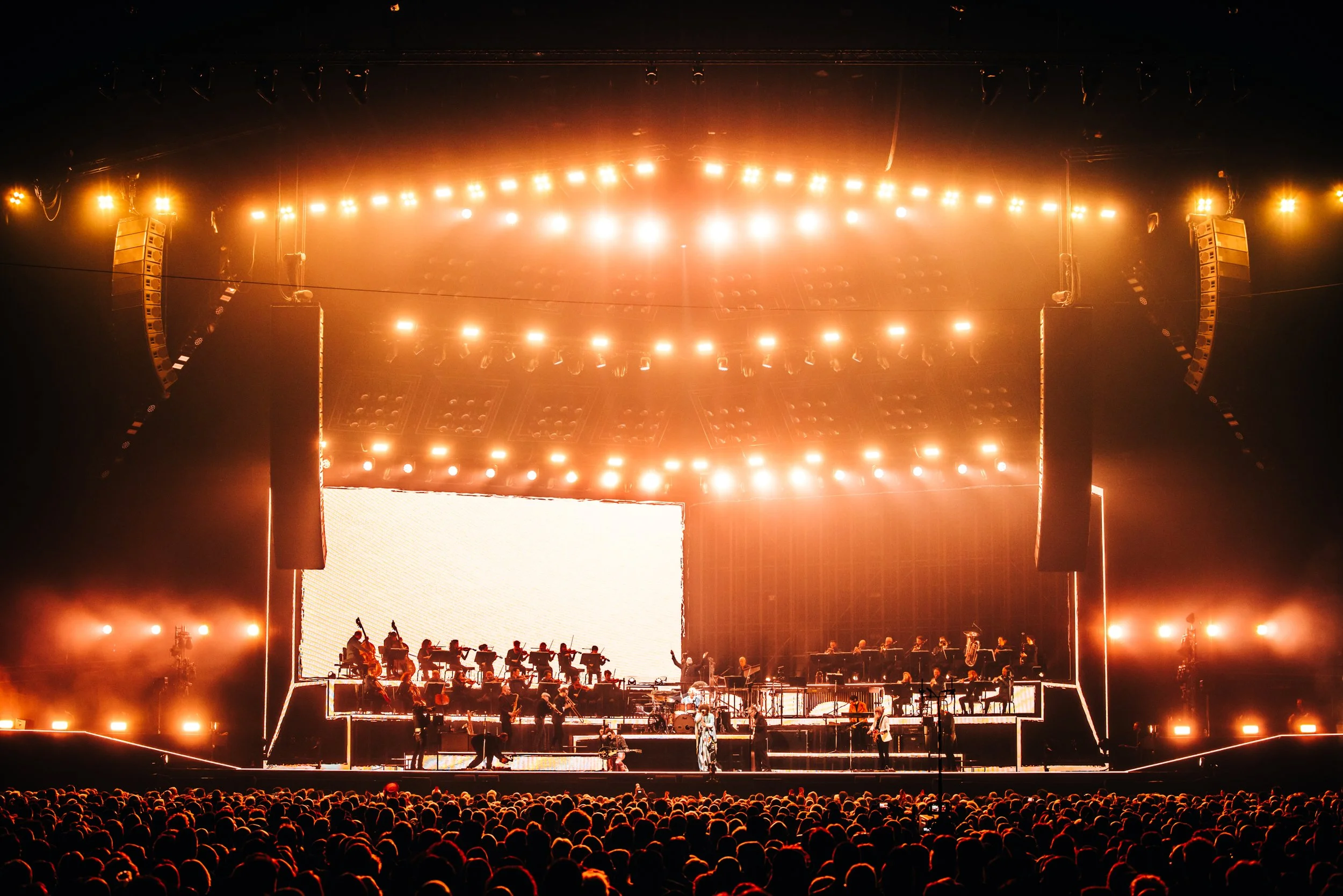 A large concert stage illuminated with bright orange and yellow lights, with musicians and crew setting up in front of a big white screen, and a large crowd gathered in front.