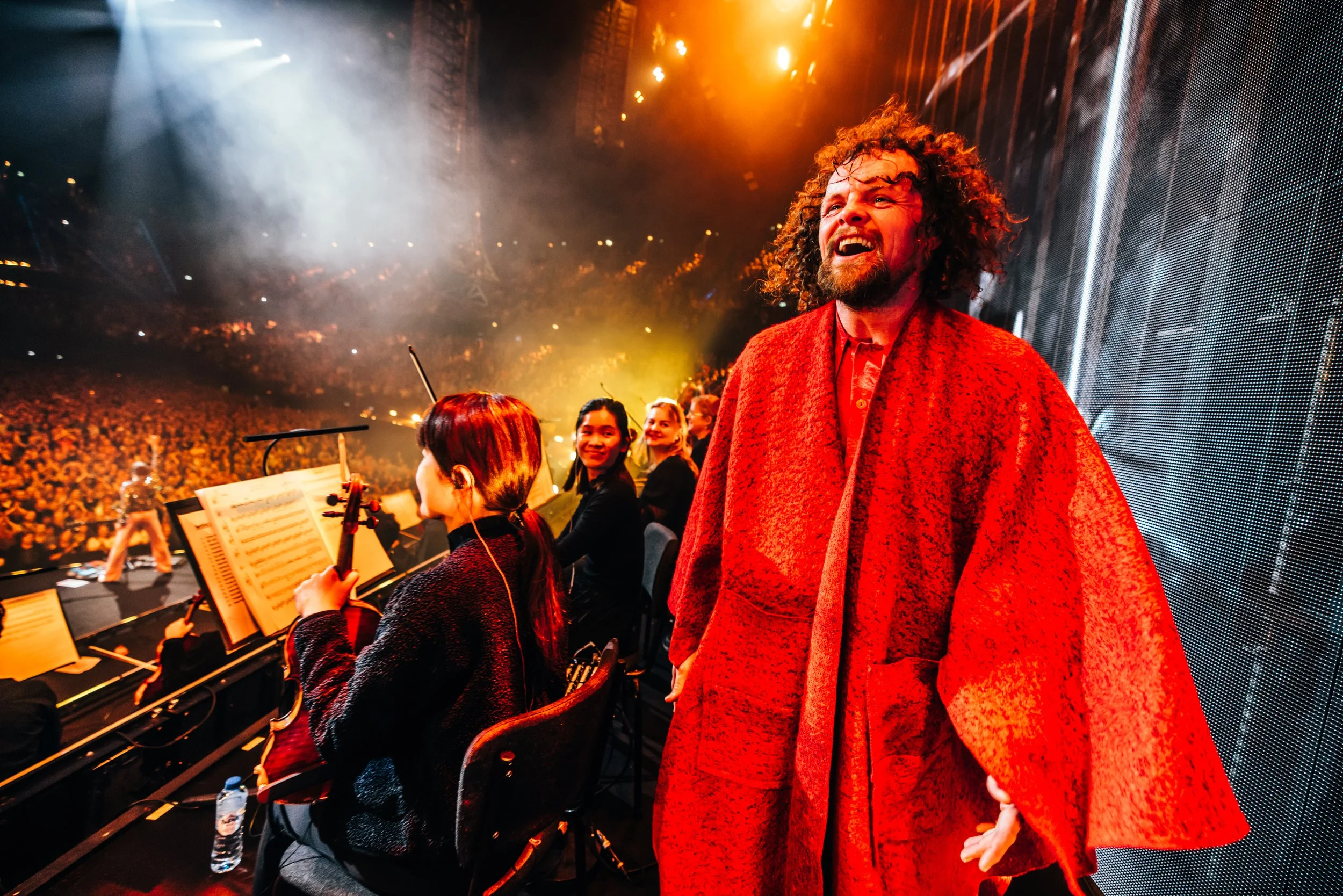 A man with curly hair and a beard wearing a red patterned coat, standing backstage at a concert. In the background, orchestra musicians and an audience in a large concert hall with warm lighting.
