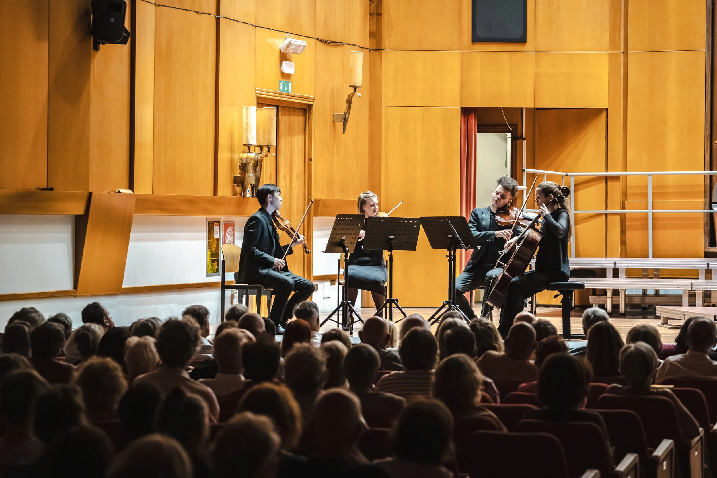 An string quartet performing on stage in front of an audience, with four musicians playing string instruments, in a wood-paneled auditorium.