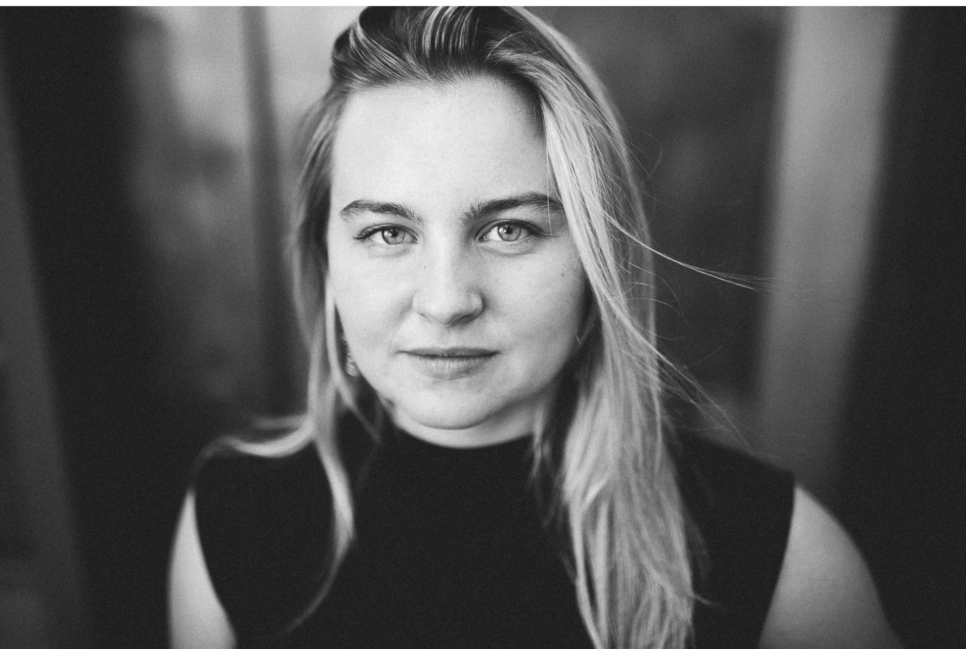 Close-up black and white portrait of a young woman with long blonde hair, looking directly at the camera with a subtle smile, wearing a dark top.