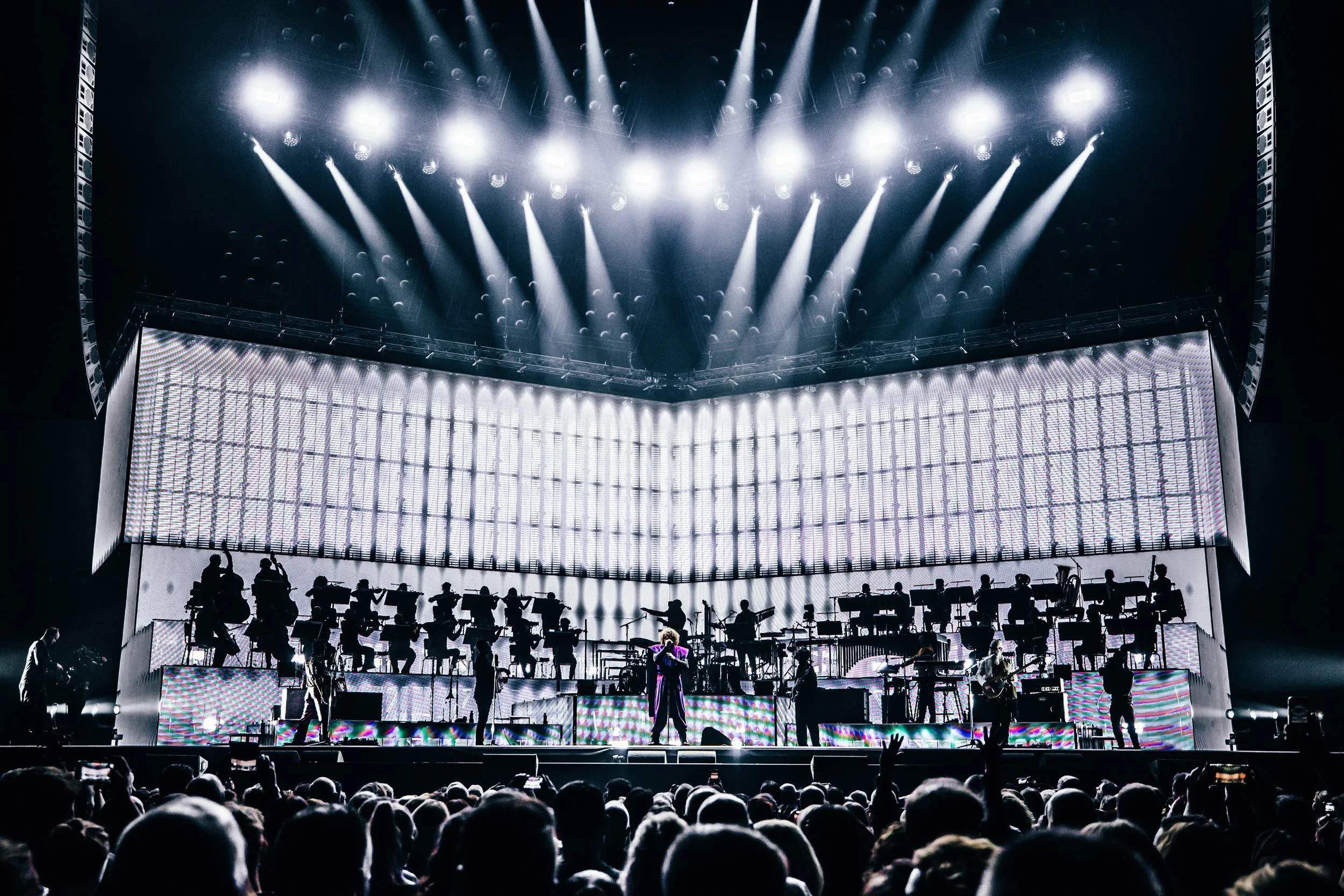 Concert stage with performers and musicians, bright stage lights, large LED screens, and an audience in front.