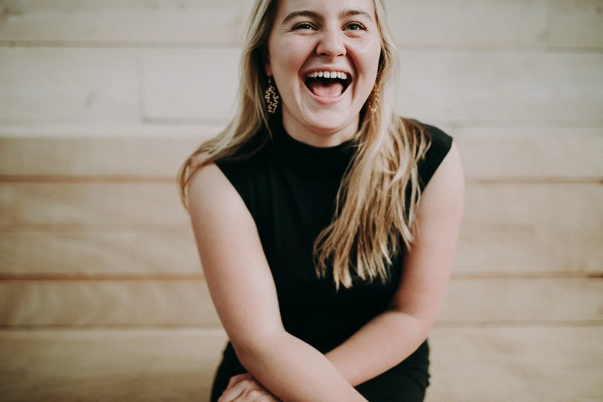 A woman with long blonde hair, wearing a sleeveless black top, is laughing with her mouth open, showing teeth, against a light wood-paneled background.