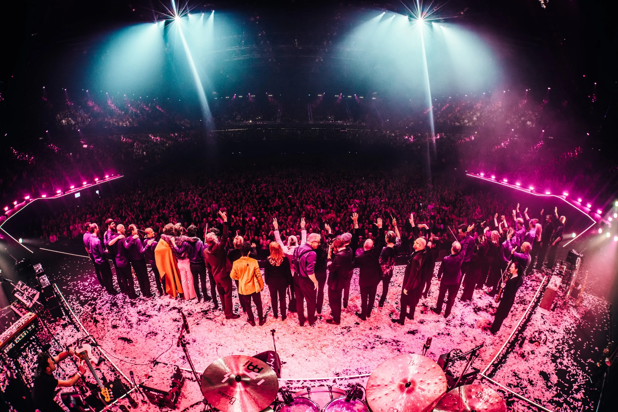 Concert scene with audience on stage, surrounded by colorful pink and blue lighting, confetti on the floor, and musical instruments including drums and guitars.
