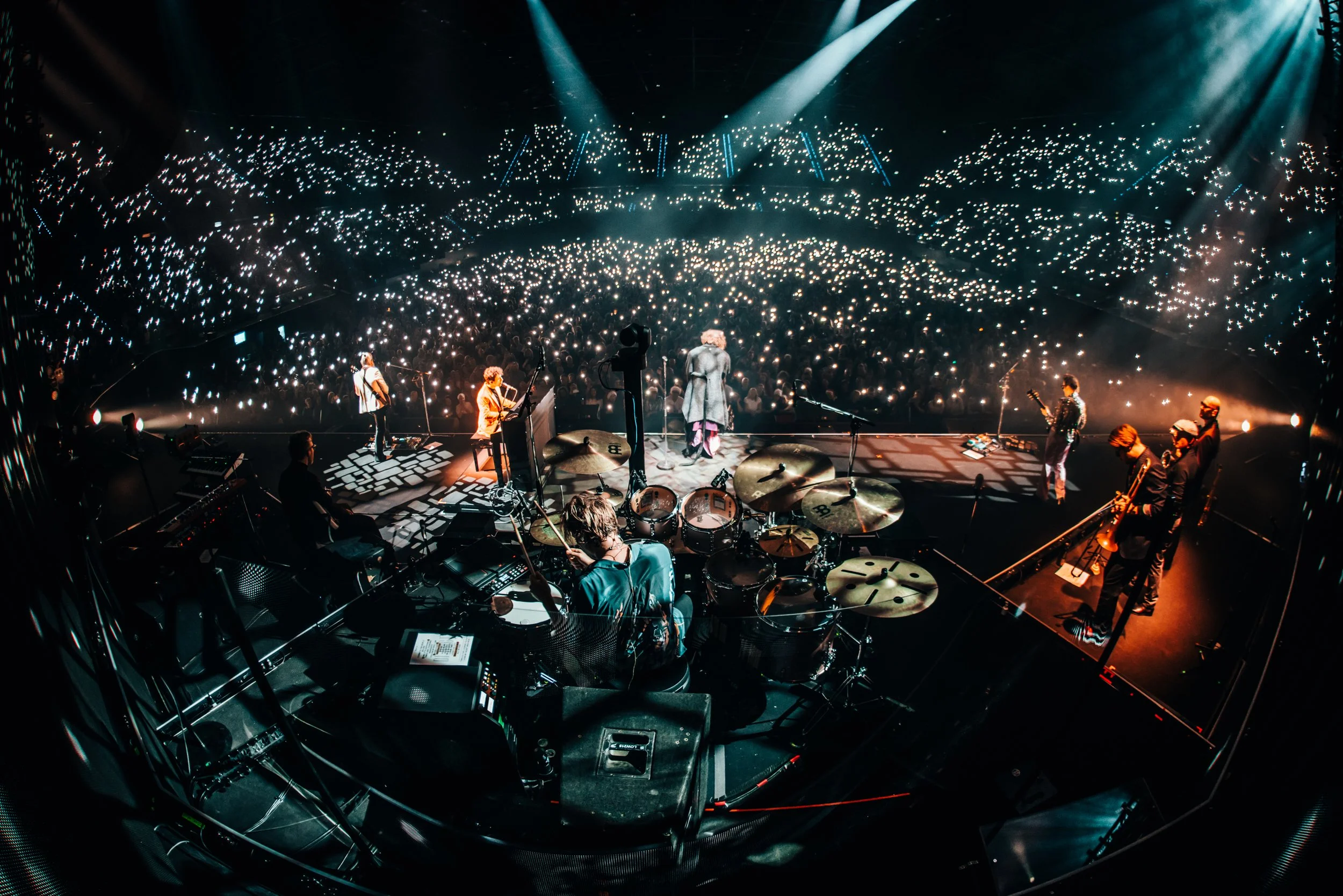 View from the stage of a live music concert, featuring musicians and a large audience holding up lights or phones with flashlights in a dark venue with spotlights overhead.