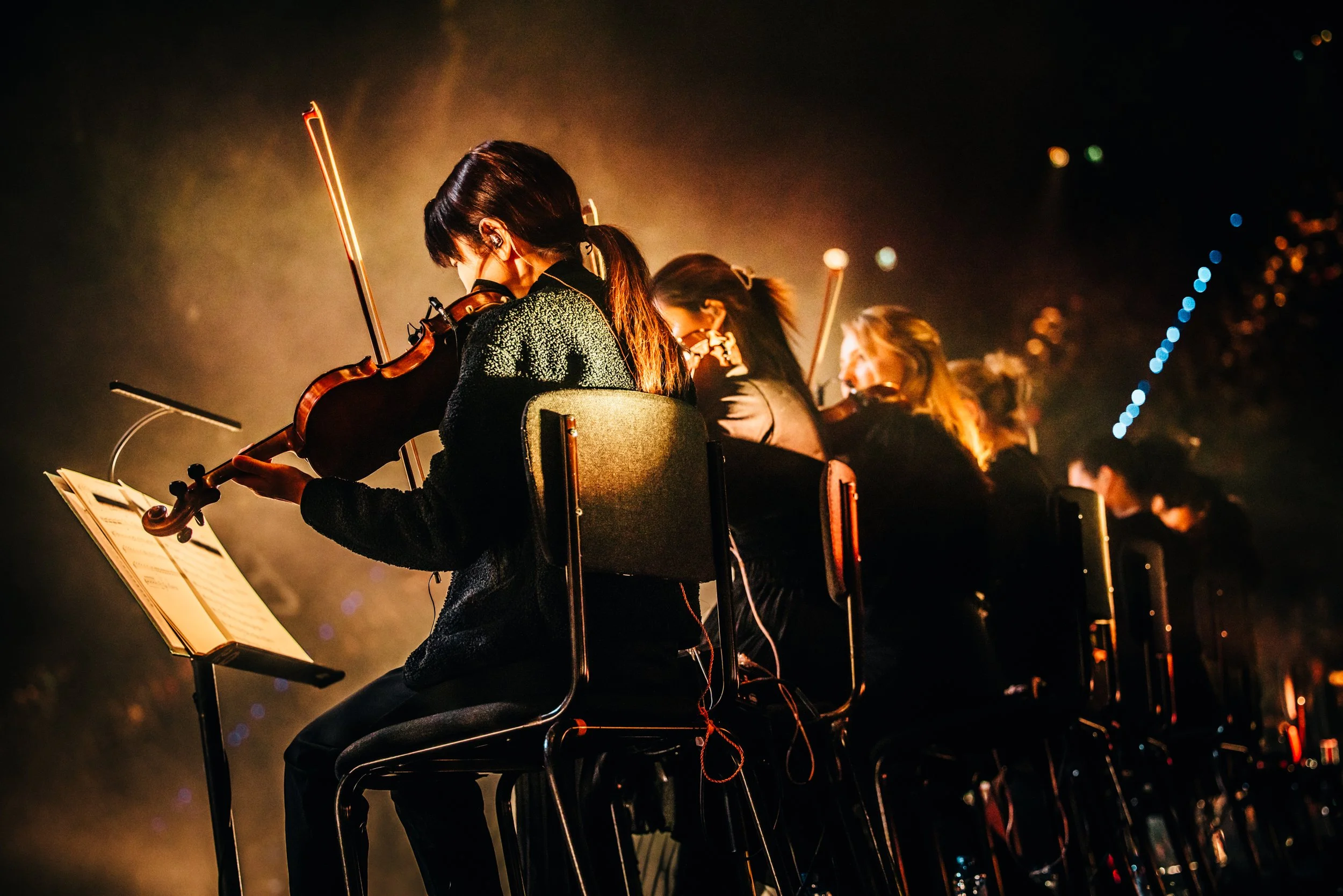 Orchestra musicians performing with DI-RECT at Rotterdam Ahoy, playing violins, with music stands and sheet music illuminated by stage lights.